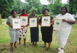 Five women stand outdoors on grass, smiling and holding framed certificates or photos. Trees and parked cars are visible in the background. The women appear to be celebrating or receiving recognition.