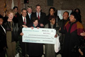 A diverse group of people smile and pose indoors while holding a large ceremonial check for $447,075 made out to Anne Arundel County, dated March 10, 2003. The event appears to be a formal celebration.