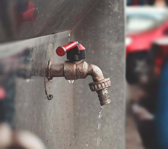 A close-up of a metal outdoor water tap with a red handle, mounted on a wall. Water is dripping slowly from the tap, and the background is blurred with hints of red and blue colors.