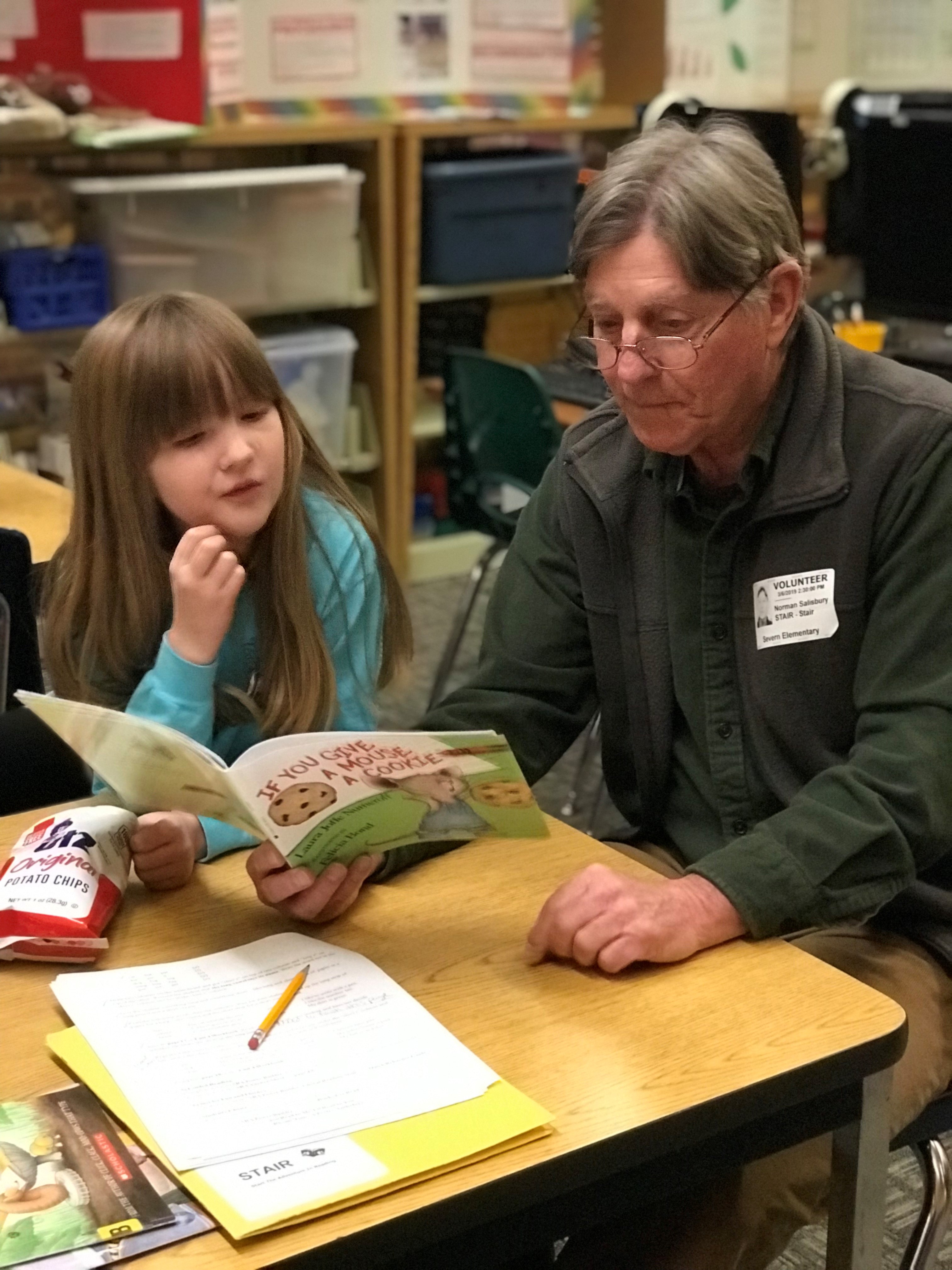 An older man with glasses helps a young girl read a book at a classroom table. Papers, a pencil, and a bag of chips are on the table. The setting appears to be a school or library.