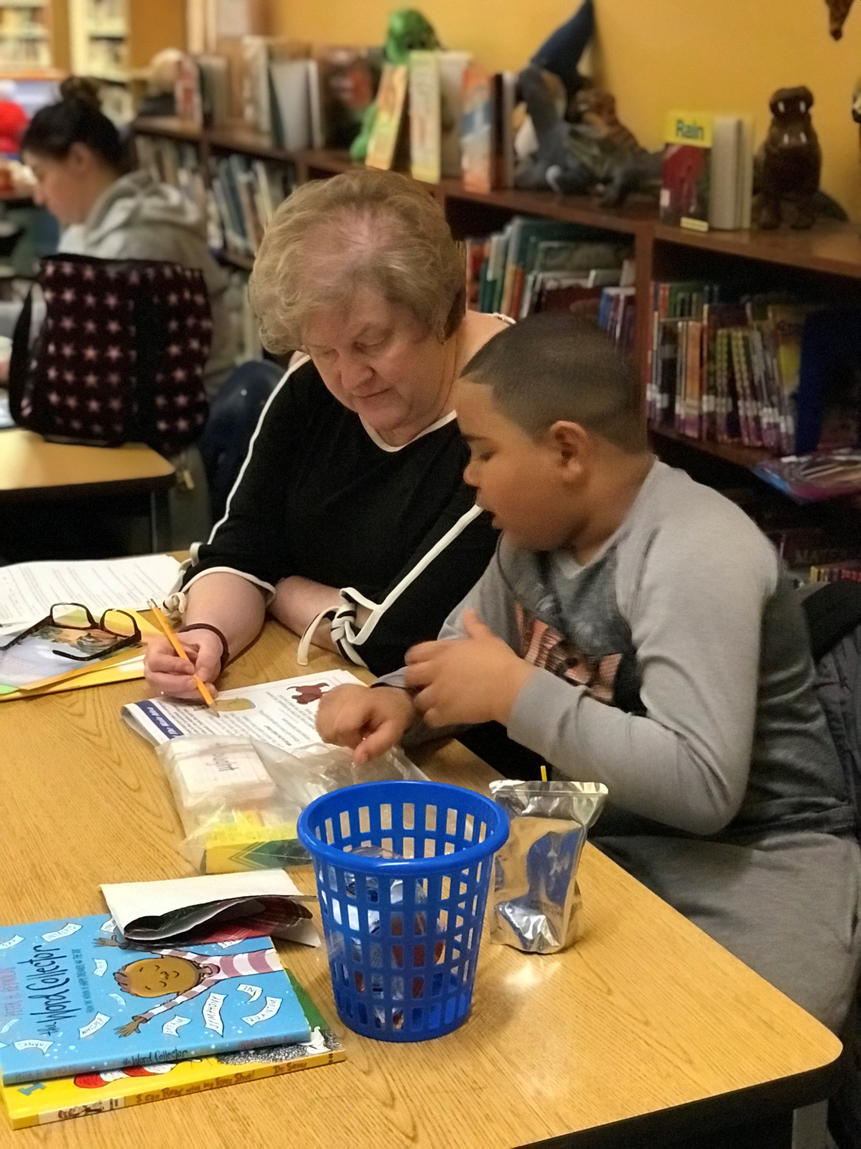 An older woman helps a young boy with schoolwork at a table in a library. They are writing together, and there are books, papers, and supplies on the table, with bookshelves in the background.