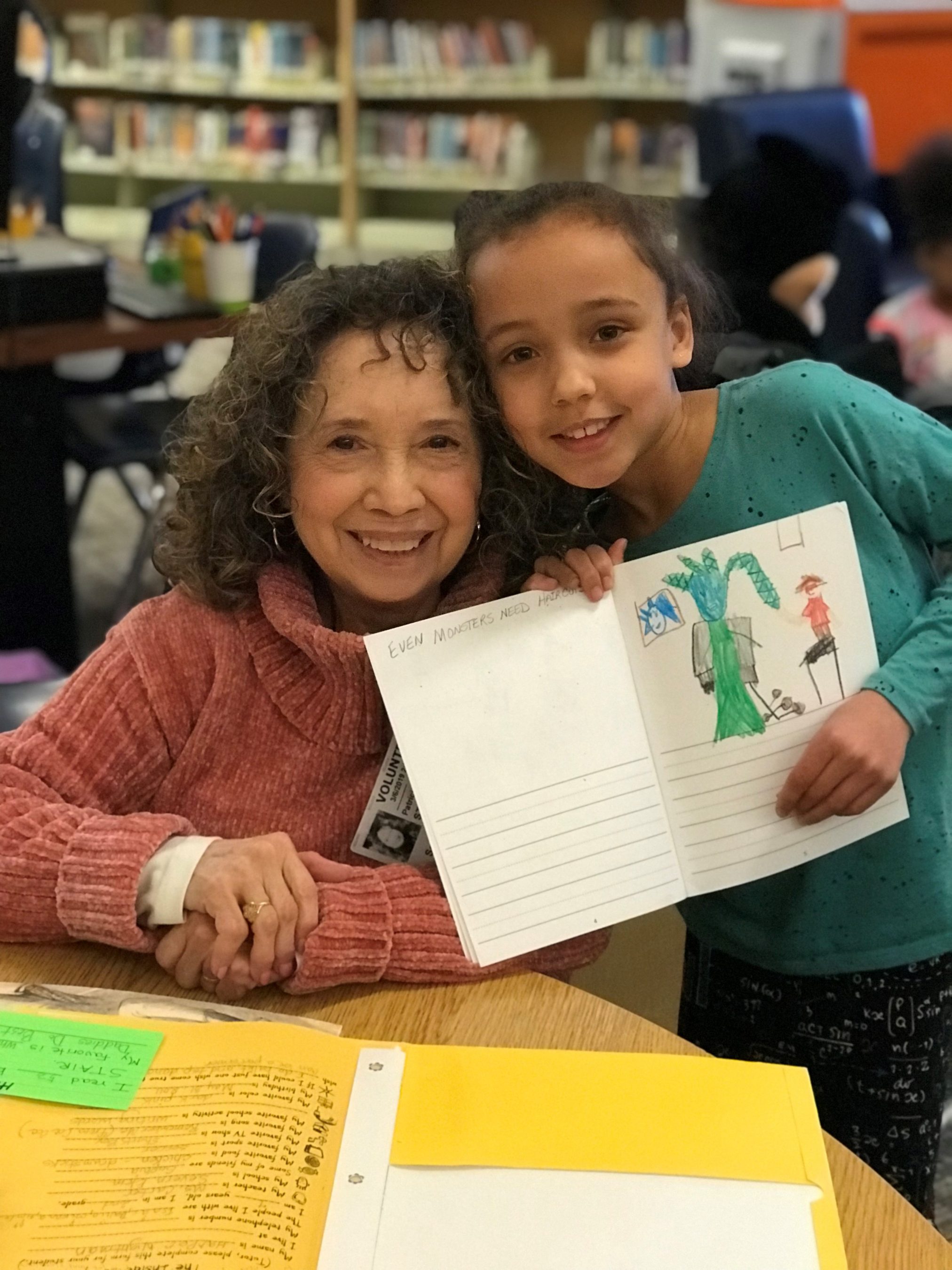 A smiling woman and a young girl pose together in a library. The girl holds up an open notebook displaying her colorful drawing and writing. Bookshelves are visible in the background.
