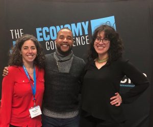 Three people stand smiling in front of a backdrop that reads "Economy Conference." Two women, one in a red blouse and one in black, stand on either side of a man wearing a gray sweater.