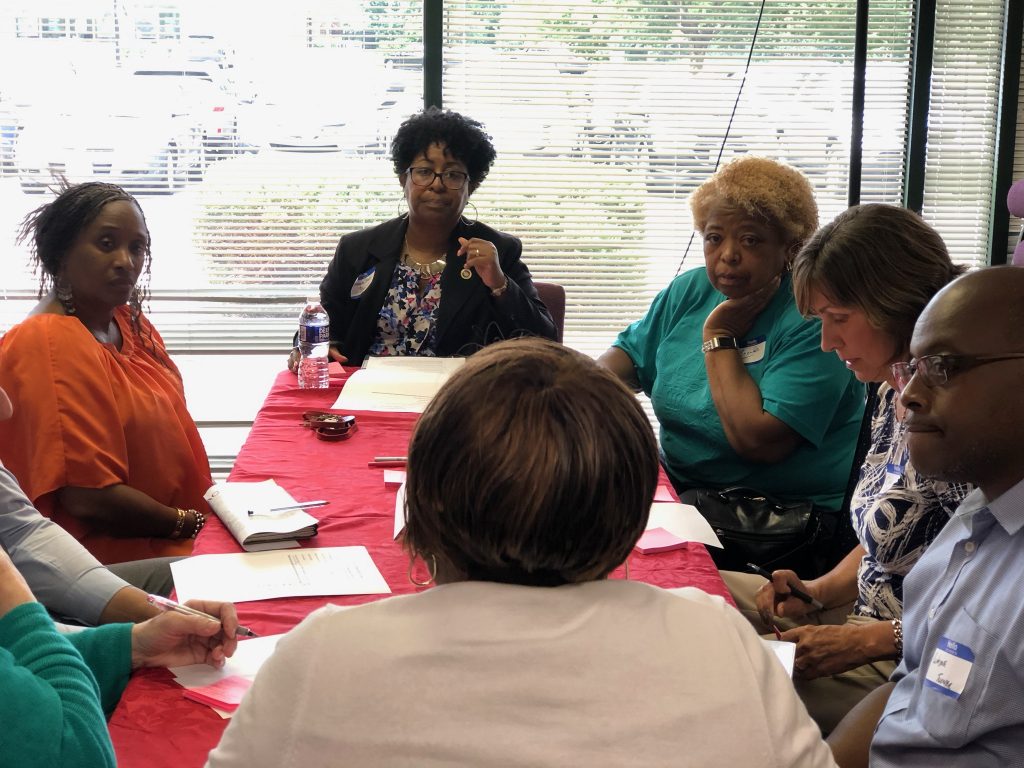 A group of seven adults sits around a table covered with a red cloth, engaged in discussion. Papers, notebooks, and pens are on the table, and large windows with blinds are in the background.