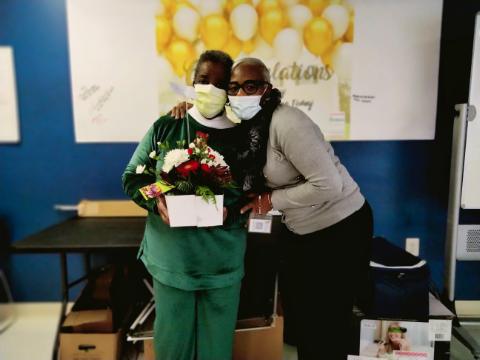 Two women wearing masks stand closely together, smiling. One holds a bouquet of flowers and a gift. There is a congratulatory banner with gold balloons in the background, suggesting a celebration or special occasion.