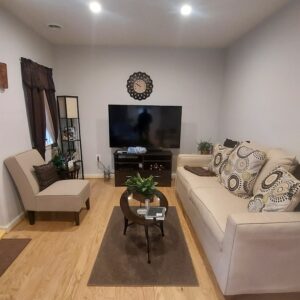 A cozy living room with a beige sofa, decorative pillows, a brown accent chair, a round coffee table, a flat-screen TV on a stand, small green plants, wooden floors, and neutral-colored walls.