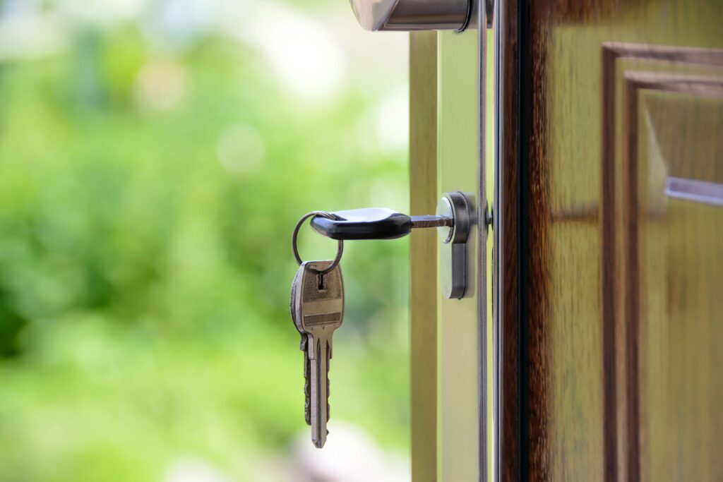 A close-up of two metallic keys on a keyring inserted into the keyhole of an open wooden door, with a blurred green outdoor background visible.
