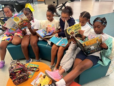 Six young girls sit together on a bench and a chair, reading colorful children’s books. They appear focused and engaged, with backpacks and more books on the table in front of them.