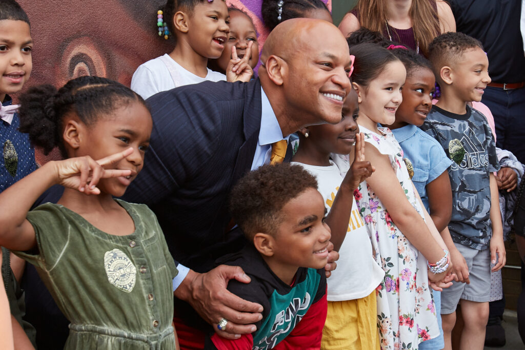 A smiling man in a suit crouches and poses for a photo with a group of happy children. The children are smiling, some flashing peace signs, and wearing colorful outfits. They are gathered closely together outdoors.