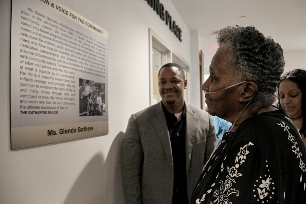 An elderly woman with an oxygen tube reads a wall plaque honoring Ms. Glenda Gathers, while two people stand beside her and smile in a hallway.