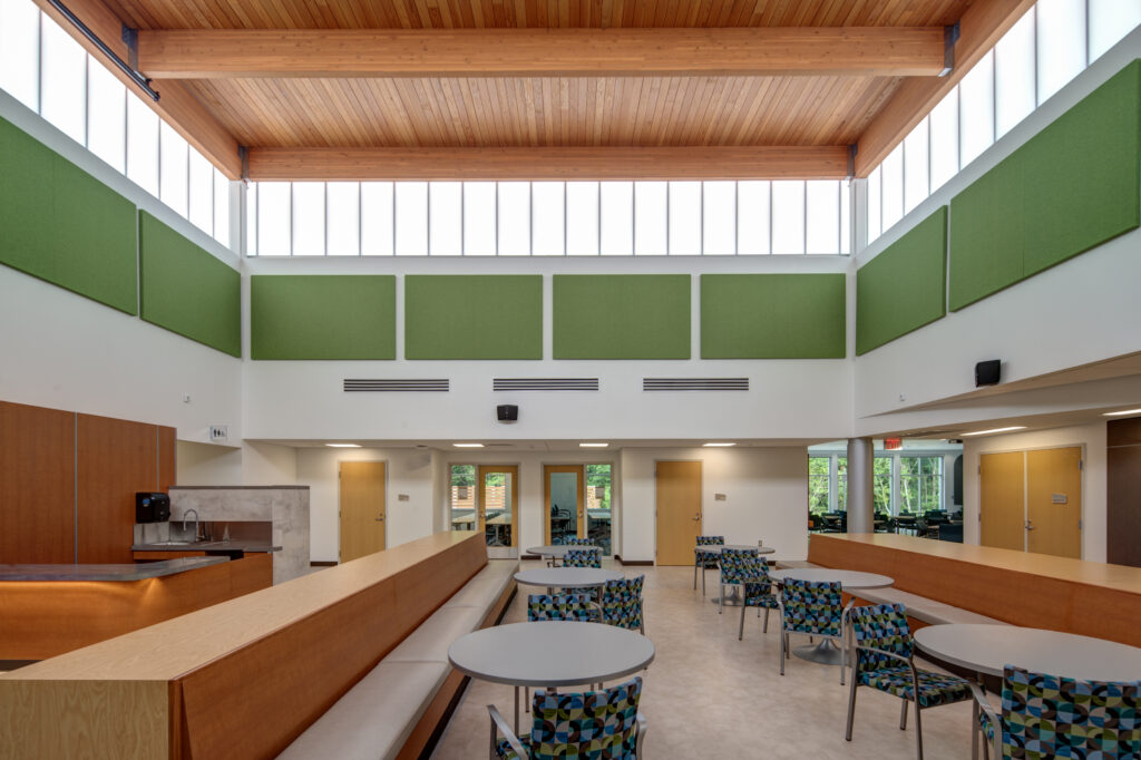 A bright, modern indoor space with high wood-paneled ceilings, large windows, green acoustic panels, round tables with patterned chairs, and a built-in wooden bench along one wall.