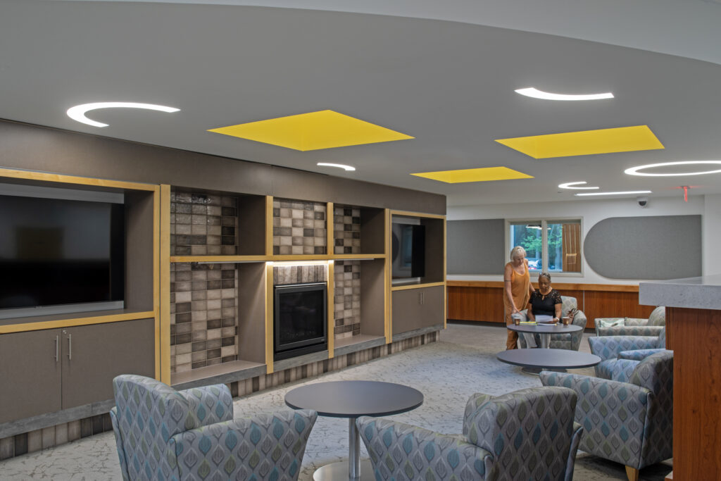 Modern lounge with patterned armchairs, round tables, a TV, built-in shelves, and a fireplace. Two people are talking by a table near large windows. The ceiling features yellow panels and circular lights.