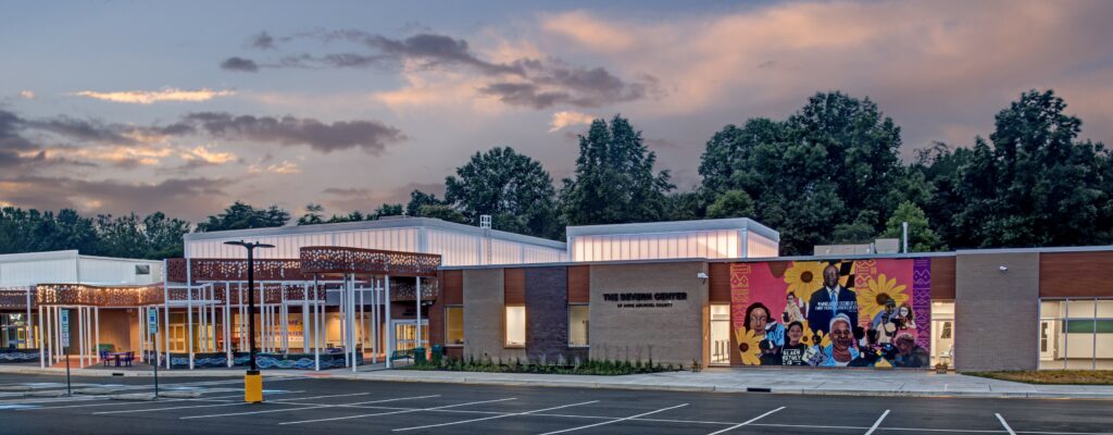 A modern school building at dusk with large windows, a vibrant mural featuring diverse people, and trees in the background. The parking lot in front is empty under a partly cloudy sky.