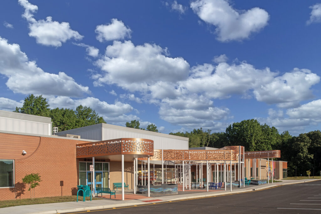 A modern school building with red brick and beige accents, featuring a covered entrance with decorative metal canopies, blue benches, and a partly cloudy sky above. Trees and a parking lot are visible in the background.