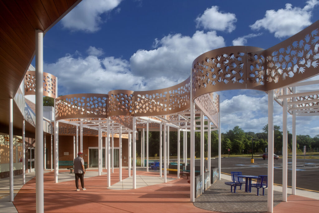 A person stands under a decorative, wavy, metal canopy with a floral cut-out pattern outside a modern building; blue sky with clouds and trees are in the background, with outdoor seating nearby.