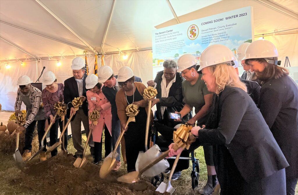 A diverse group of people in hard hats stand under a tent, holding shovels with gold bows, breaking ground for a new project. A large sign in the background announces "Coming Soon Winter 2023.