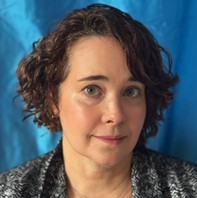 A woman with short, curly brown hair and light skin looks at the camera against a blue background. She is wearing a patterned top and has a neutral expression.