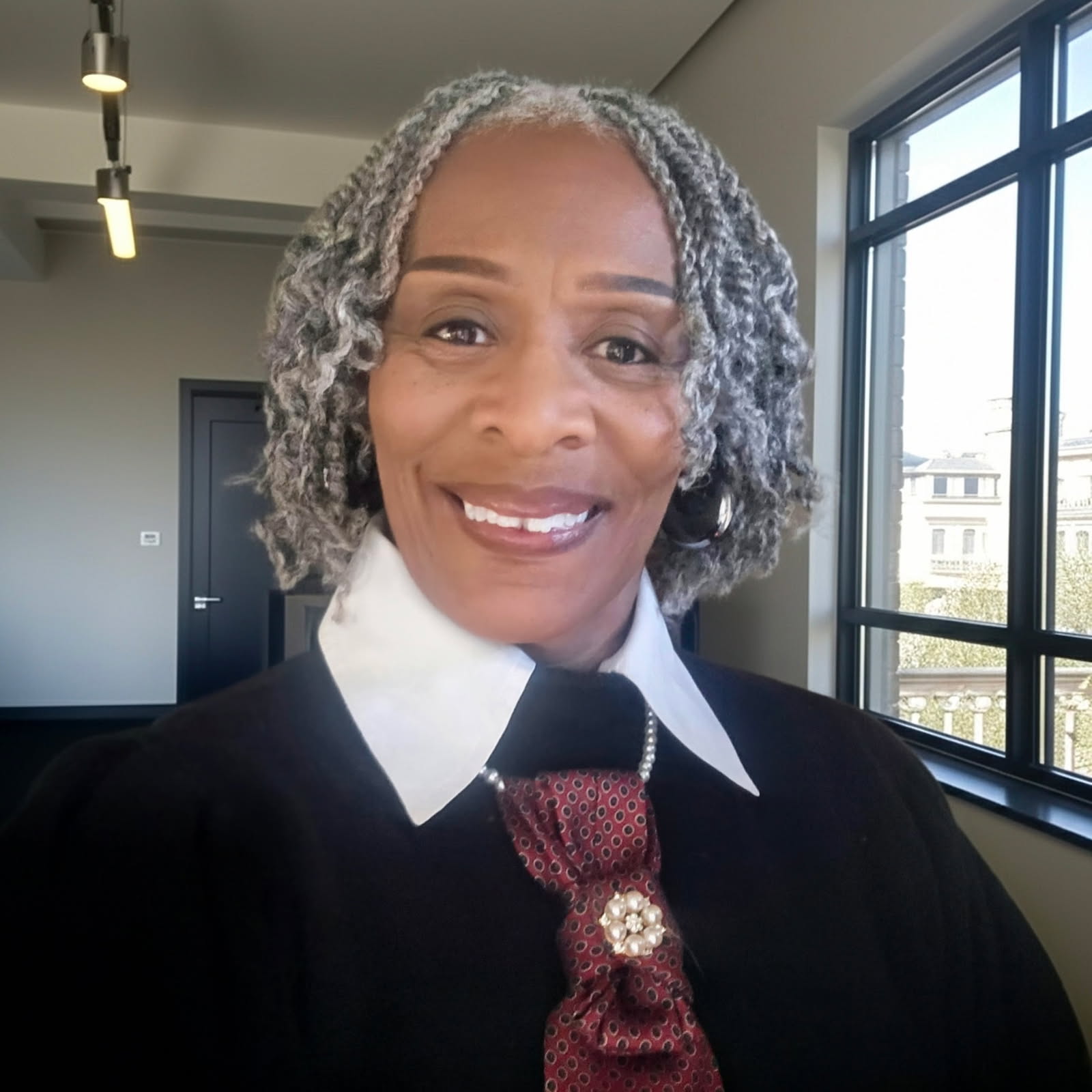 A smiling older woman with gray curly hair wears a white-collared shirt, black sweater, and a decorative red tie. She stands indoors near large windows overlooking a cityscape.